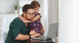 Dad looking at his computer while putting his arm around his daughter while she watches